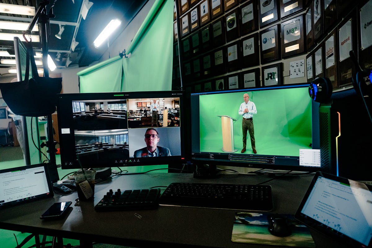 A computer station shows a video call on one monitor and a man in front of a green screen on another monitor.