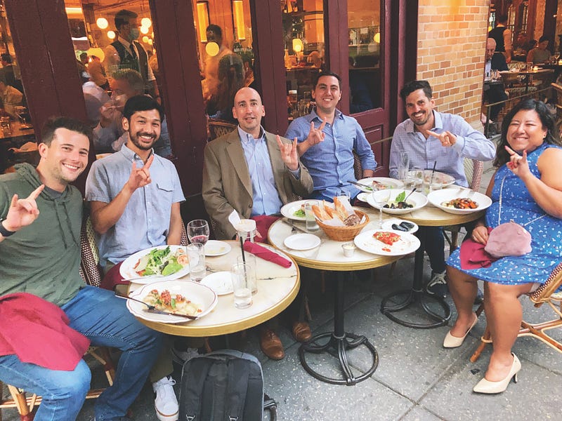 Group of casually dressed McCombs alumni using Hook ’em Horns hand sign outside at a restaurant.