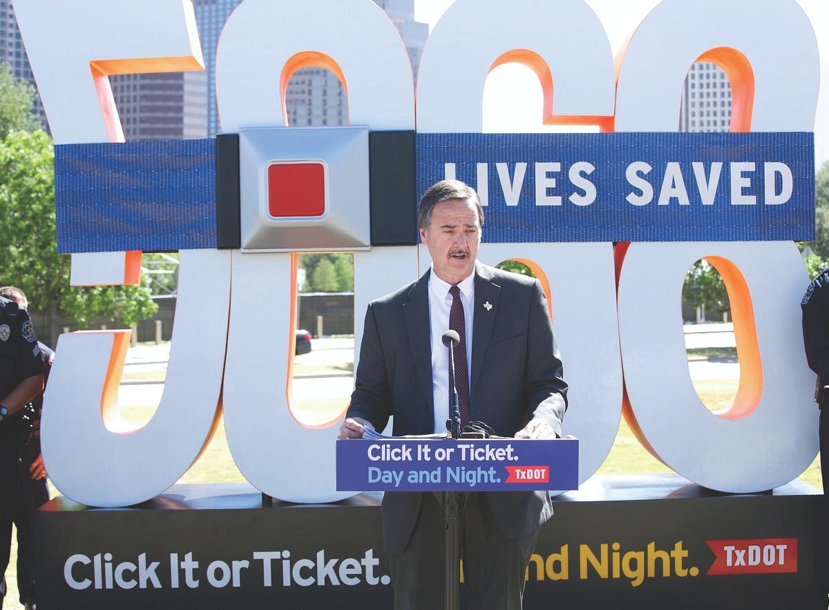 A man in a black suit speaks in front of a sign that reads “5000 Lives Saved. Click It or Ticket. Day and Night.”