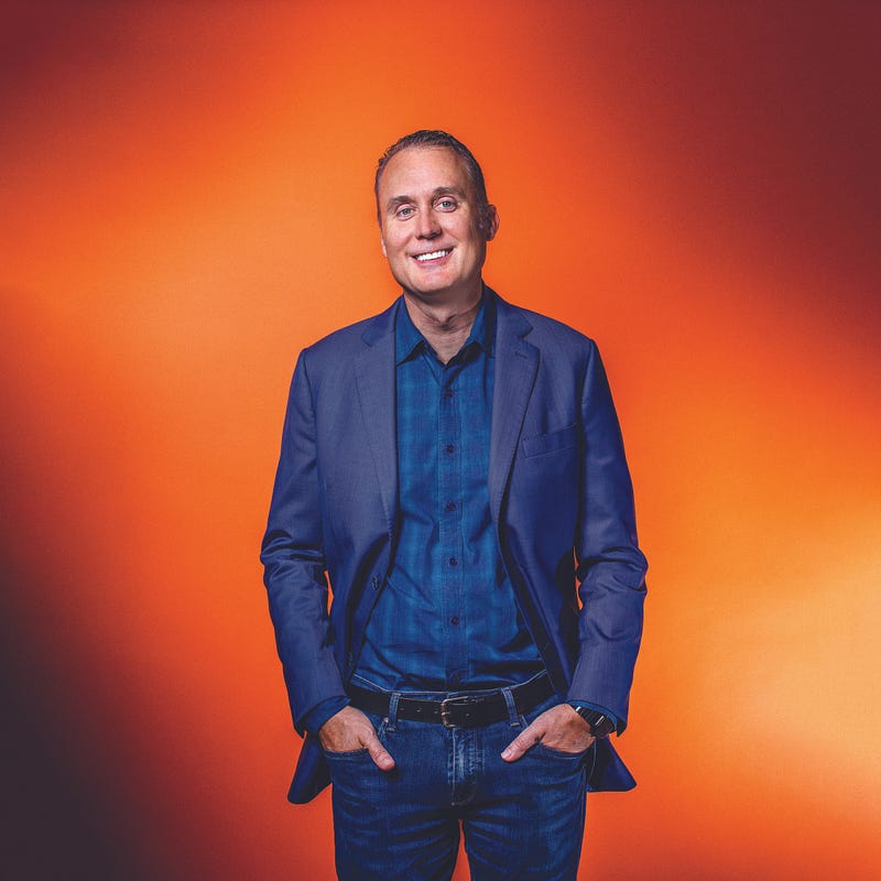 Brett Hurt smiles wearing a blue patterned shirt, jeans, and a navy jacket against an orange background.
