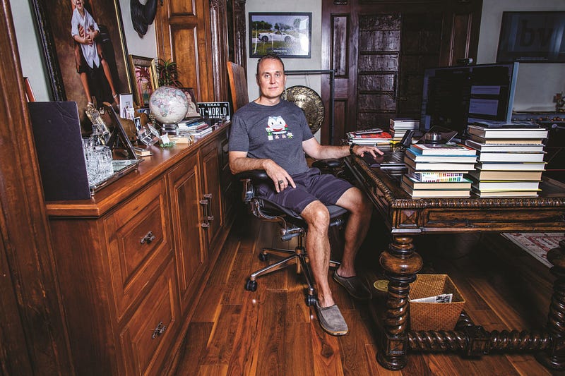 Brett Hurt sits in his home office behind a desk with a monitor and stacks of books.