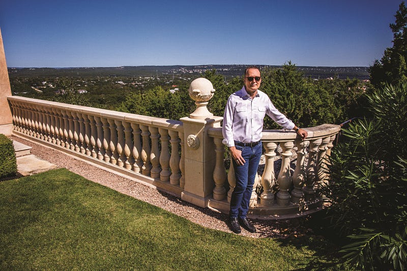 Brett Hurt stands in front of stone fence circling his house in Austin, with a view of hills in the background.