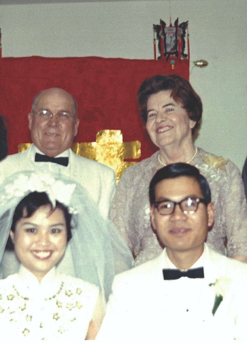A young woman in a wedding dress sits next to a young man in a white tuxedo, with an older couple standing behind them.