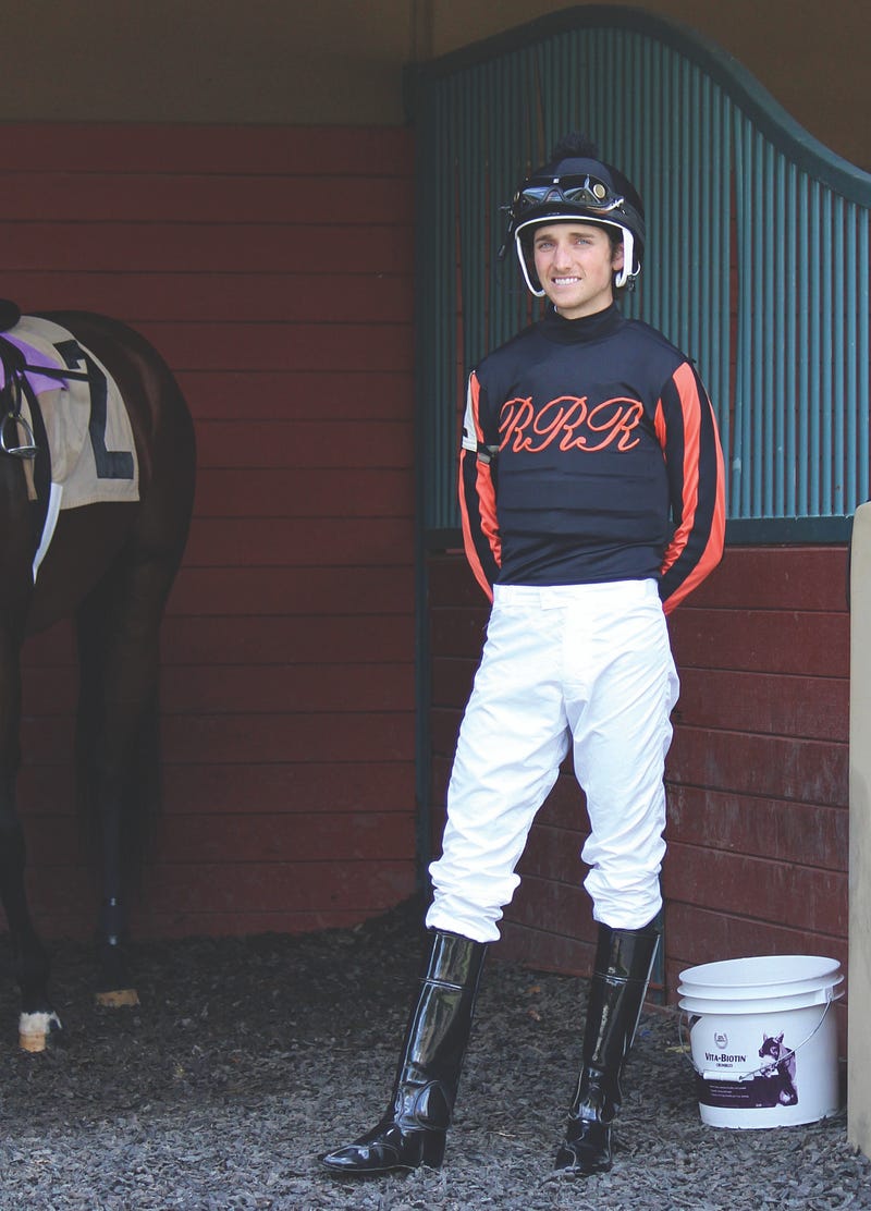 A uniformed male jockey stands in a stable next to his horse.