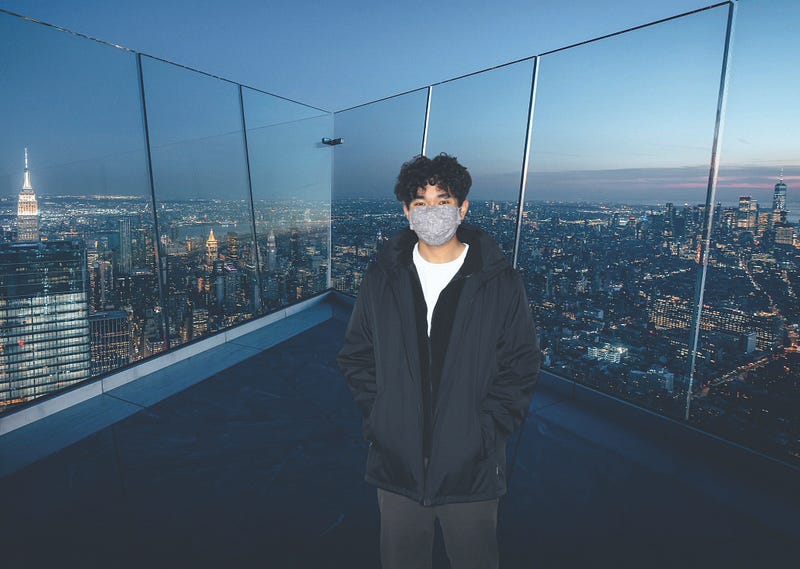 A man with curly hair and a face mask stands on top of the Empire State Building with the New York City skyline behind him.