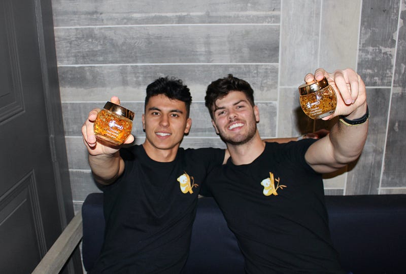 Two men hold small, orange, glass jars to the camera