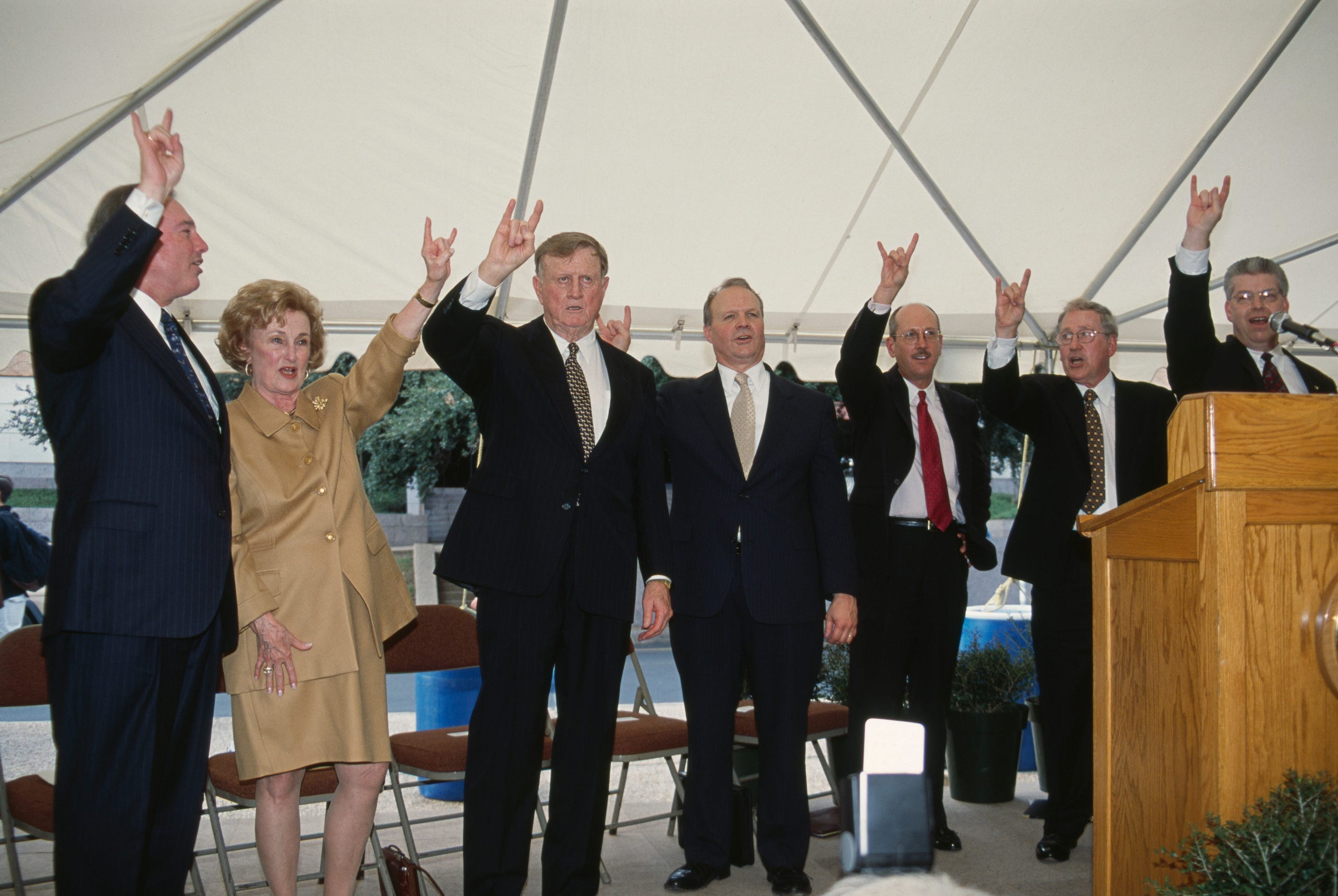 Red McCombs, Charline McCombs, and University of Texas staff stand on a stage making the Hook ’em Horns hand symbol.