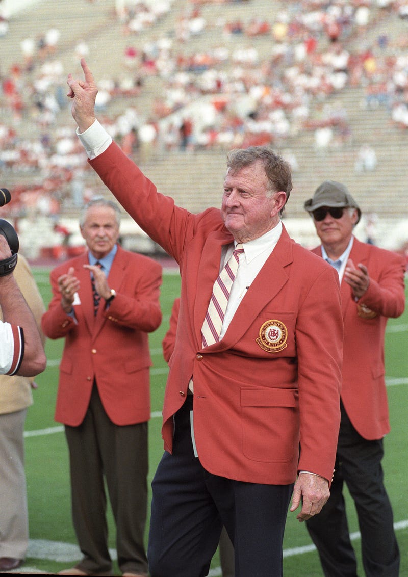 Red McCombs waves to the crowd on a football field, with men behind him clapping.