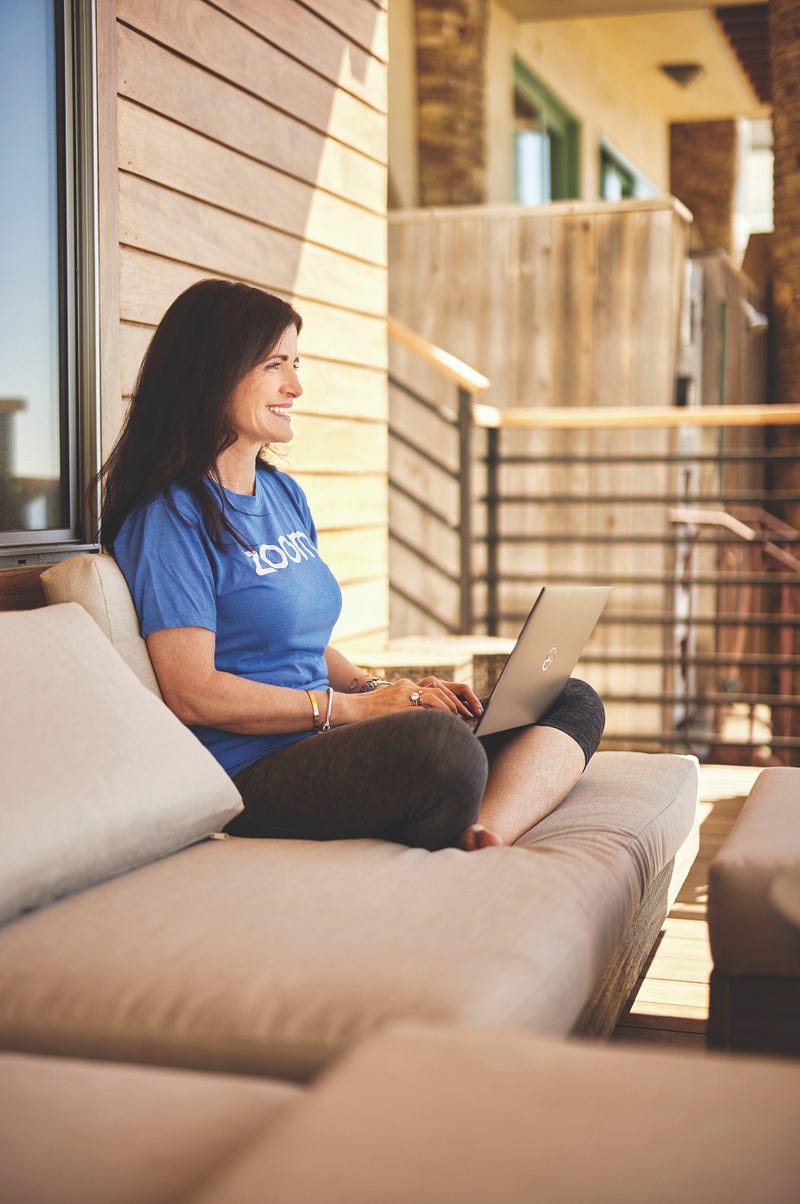 Kelley Steckelberg wears a Zoom t-shirt and sits cross-legged on an outdoor couch while typing on her laptop.