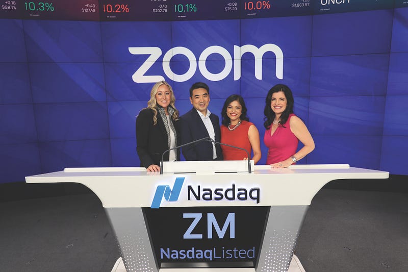 Zoom executives stand behind a desk at the Nasdaq stock market with a screen showing Zoom’s logo in the background.