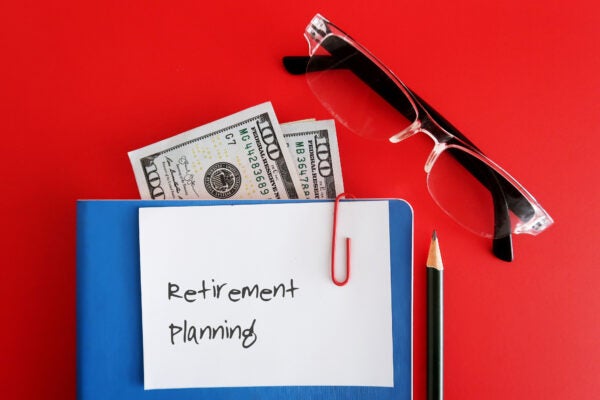 A note written RETIREMENT PLANNING clip with a blue notebook,cash dollar banknote inside, with a pencil and a reading glasses, isolated on red background. Concept of financial planning for retirement security