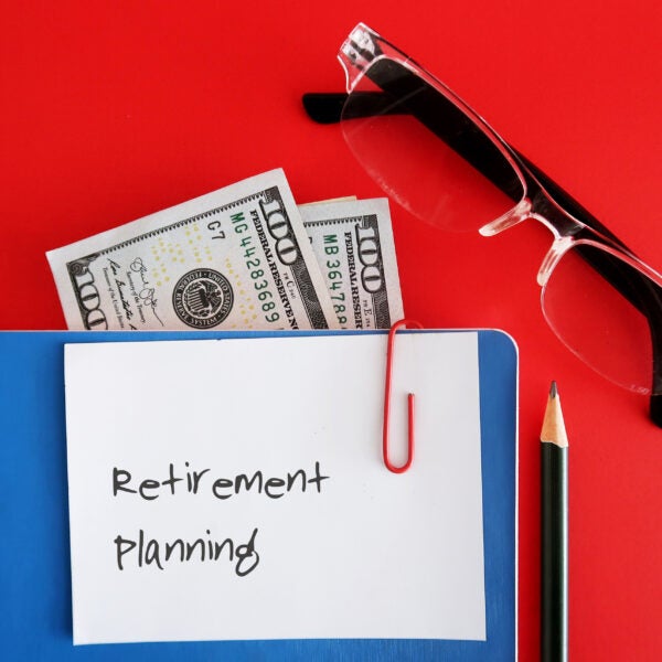 A note written RETIREMENT PLANNING clip with a blue notebook,cash dollar banknote inside, with a pencil and a reading glasses, isolated on red background. Concept of financial planning for retirement security